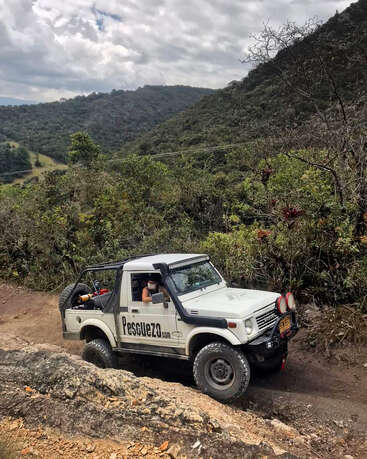 A rugged white off-road vehicle navigates a rocky dirt trail, surrounded by dense greenery and mountainous landscape under a cloudy sky. Adventure and exploration are evident.