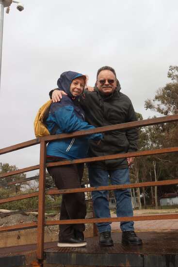 A smiling couple in jackets stands on a rusty metal bridge outdoors. The woman wears a blue raincoat and backpack, while the man wears sunglasses and smiles.