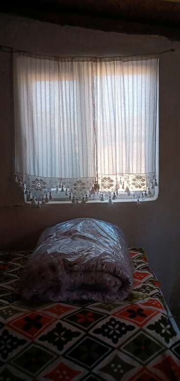 A soft light filters through sheer, lace-trimmed curtains. Below, a plastic-wrapped folded blanket rests on a bedspread with a colorful geometric pattern. Cozy atmosphere.