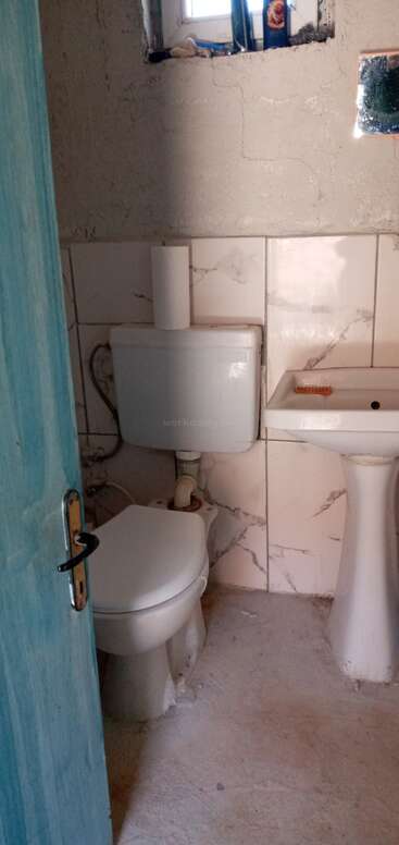 This image shows a small bathroom with a white toilet, a pedestal sink, a blue door, unfinished walls, a small window, and a mirror.