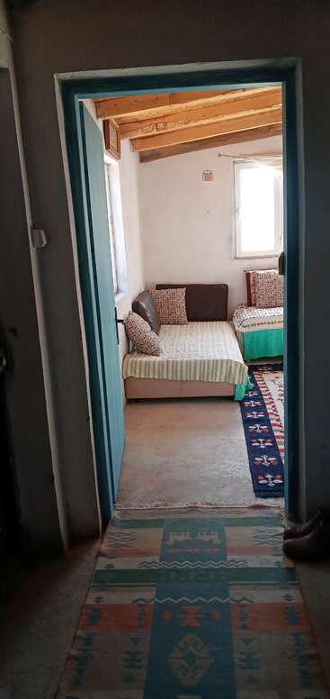 This image shows a cozy, rustic room with wooden ceiling beams, patterned rugs, simple beds with cushions, and sunlight coming through a window, creating a warm atmosphere.