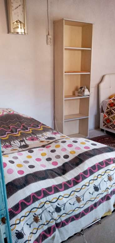 This image shows a cozy bedroom with a patterned bedspread, a wooden bookshelf, an iron, a wall clock, and a white bed in the corner.