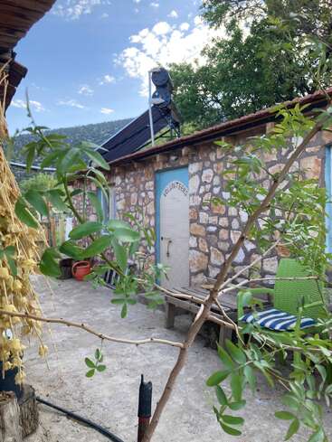 Um prédio rústico de pedra com a placa “Voluntários” tem um aquecedor solar de água no telhado. Cercado por vegetação, bancos ao ar livre e ferramentas de jardinagem, com as montanhas ao fundo.
