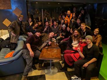 The image depicts a group of approximately 20 young adults gathered in a dimly lit room, posing for a photo with a pool table and couch in the background.