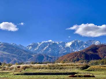The image depicts a serene mountainous landscape with a grassy field in the foreground, featuring a house and vibrant autumn foliage, set against a clear blue sky.