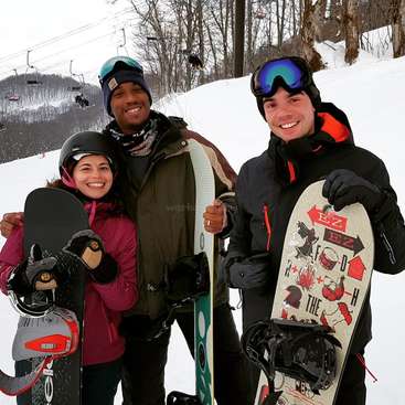 The image shows three people posing for a photo on a snowy hill, each holding a snowboard and wearing winter gear. They are smiling at the camera.