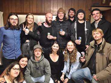 The image depicts a group of 12 young adults posing for a photo, with some holding drinks, in a cozy room with wood paneling on the walls.