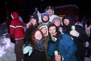 A group of friends huddles together outdoors at night in snowy winterwear, smiling and holding drinks, enjoying a fun, festive evening in the snow.