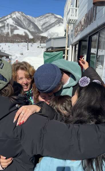 A group of friends share a warm group hug outdoors in a snowy mountain setting, with bright sunlight, laughter, and visible joy radiating from everyone.