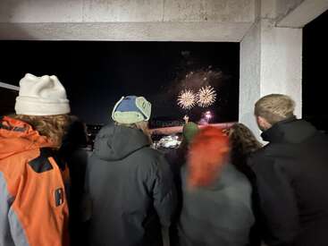 A group of people dressed in winter clothes watch colorful fireworks explode in the night sky, framed by a concrete structure, capturing a festive celebration.