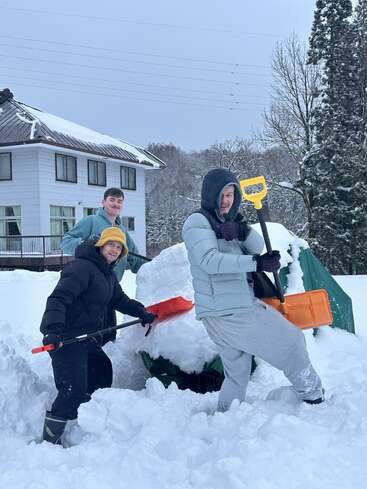 Three people bundled in winter clothes are smiling while building an igloo in the snow with shovels. A snowy house and trees are in the background.