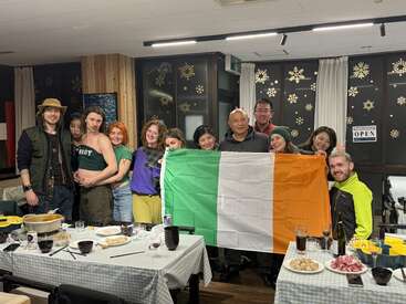 A group of people smiles together indoors, holding an Irish flag. Festive decorations and food on tables suggest a joyful gathering or celebration in a cozy setting.