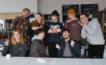 Eight people pose playfully indoors, making heart signs and funny gestures. They appear cheerful and relaxed, sitting and standing together, in a cozy, casual setting with menus behind.