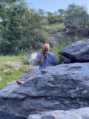 Un homme est assis sur de gros rochers à l'extérieur, en train de parler au téléphone. Il est pieds nus, porte une chemise à motifs et se trouve au milieu de la verdure, par une journée ensoleillée.