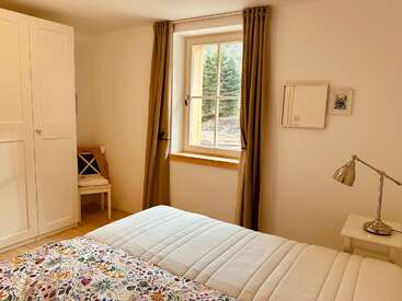 This cozy bedroom features a large window with brown curtains, a white wardrobe, a chair with cushions, a lamp, and a bed with floral bedding.
