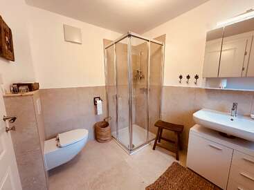 This bathroom features a glass shower, wall-mounted toilet, modern sink cabinet, wooden stool, mirror cabinet, basket, hooks, beige tiles, and minimalistic decor.
