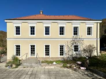 A two-story beige building with a red-tiled roof, white trim, and symmetrical windows stands under a clear blue sky, surrounded by greenery and steps.