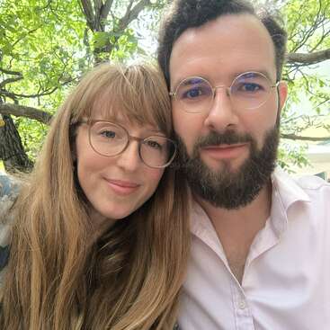 A couple poses closely together outdoors, both wearing glasses and smiling softly. Green leaves and tree branches create a natural, serene background, suggesting a peaceful, happy moment.