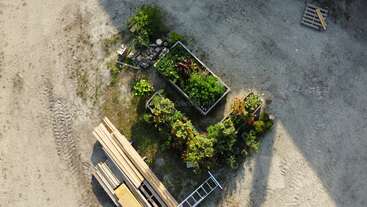 This aerial photo shows a small garden with raised beds and green plants, surrounded by dirt, building materials, wooden planks, a ladder, and scattered pallets.