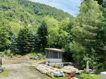 A small wooden shed sits near piles of lumber and scattered construction materials, surrounded by lush green trees and hills under a bright, clear blue sky.