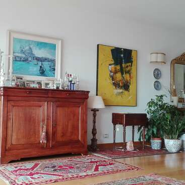 La imagen muestra un salón con un gran ventanal, en el que hay un sofá y una mesa de centro, y una chimenea, con una estantería y objetos decorativos en las paredes.