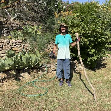 Un hombre está de pie en un jardín, sosteniendo un palo grande y una herramienta eléctrica. Sonríe, lleva un sombrero, guantes y ropa informal, y está rodeado de plantas.