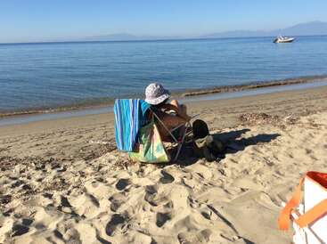 Una persona se relaja en una silla en una playa de arena, frente al mar en calma, con un sombrero. Un barco flota en la distancia bajo un cielo despejado.