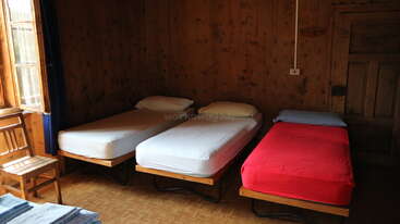 The image shows a room with three beds, each with a mattress and bedding, and a table and chairs in the background, likely a dormitory or hostel.