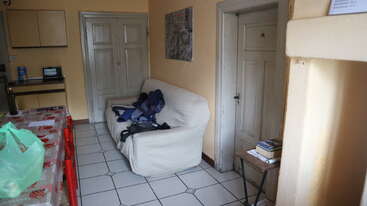 The image depicts a living room with a couch, coffee table, and TV, featuring a large window and a fireplace, with a bookshelf and a doorway visible in the background.