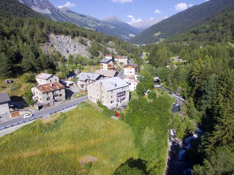 The image depicts a serene mountain village, featuring a cluster of buildings amidst lush greenery and a winding road, set against a picturesque backdrop of mountains and a blue sky.