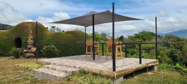 A serene outdoor patio features two wooden chairs and a small table under a shade sail, overlooking lush greenery, with a grass-covered structure and stone sculpture nearby.