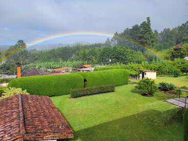 A vibrant rainbow arches across a lush, green landscape with neatly trimmed hedges, trees, countryside buildings, and a peaceful, cloudy sky enhancing the picturesque scene.