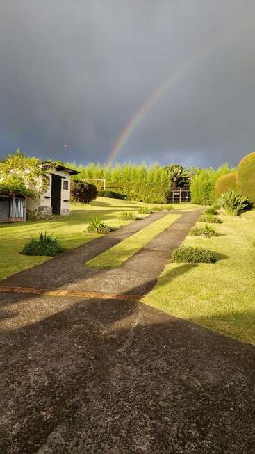 A stone driveway leads to a lush garden and a small shed. Overhead, dark clouds contrast with a vibrant rainbow arching across the sky.