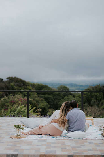 A couple sits closely on a blanket outdoors, enjoying a peaceful, romantic moment together. They overlook lush greenery and cloudy skies, embracing each other lovingly.