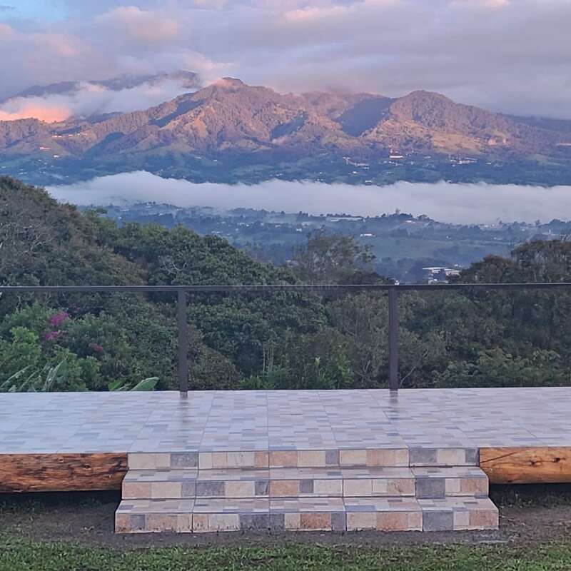 The image depicts a serene outdoor setting, featuring a wooden structure with a flat roof, surrounded by lush greenery and a body of water, evoking a sense of tranquility and natural beauty.