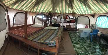 Cette image montre l'intérieur d'une tente spacieuse avec deux lits, une literie colorée, plusieurs étagères de rangement, une table, des chaises en plastique et un tapis de sol à motifs, le tout sous une douce lumière du jour.