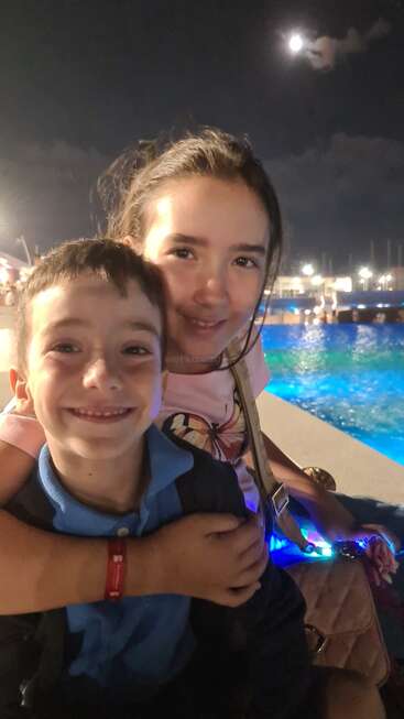 Dos niños sonrientes posan para una foto de noche, iluminados por luces brillantes cerca de una piscina. La luna brilla en el cielo, creando un ambiente cálido y alegre.