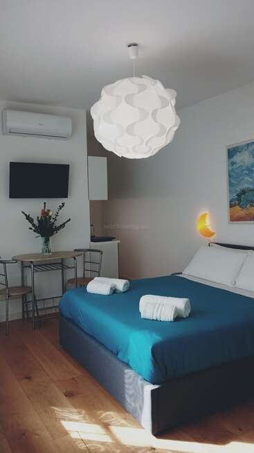 Un acogedor dormitorio cuenta con una cama revestida de color verde azulado, una moderna lámpara colgante, un aplique de media luna amarillo, una pequeña mesa de comedor, un televisor, aire acondicionado, obras de arte y suelo de madera.