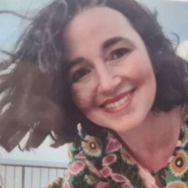 Una mujer sonriente con el pelo corto, ondulado y oscuro lleva un colorido vestido estampado. Está inclinada hacia delante, al aire libre, con un fondo de cielo azul pálido.