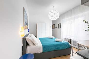 Dormitorio moderno y luminoso con colcha verde azulado, dos toallas enrolladas, armario blanco, ventana grande con cortinas transparentes, lámpara colgante decorativa, obras de arte y mesa de comedor pequeña.