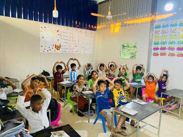 A classroom filled with cheerful children sitting at desks, making heart shapes with their arms above their heads. Colorful posters and educational materials decorate the walls.
