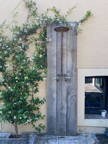 Das Bild zeigt eine Außendusche mit einer rustikalen Holzverkleidung, Metallarmaturen und einer Kletterjasminpflanze mit weißen Blüten neben einem Fenster an einer beigen Wand.