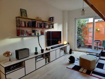 This modern living room features minimalist furniture, a TV, bookshelves, and a black cat. Large windows open to a patio with orange walls and greenery.