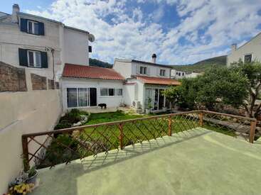 Dieses Bild zeigt einen Hinterhof mit grünem Gras, einen kleinen Garten, einen schwarzen Hund, weiße Häuser mit roten Dächern, Bäume, blauen Himmel und vereinzelte Wolken.