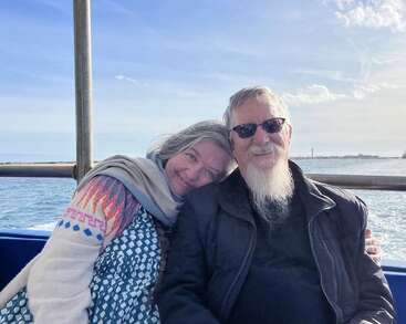 Una pareja de ancianos disfruta de un soleado paseo en barco. La mujer se apoya cariñosamente en el hombro del hombre, ambos sonrientes, rodeados de agua y cielo azules.