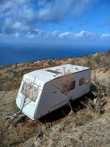 Ein weißer Wohnwagen steht auf einem trockenen, grasbewachsenen Hügel mit Blick auf den Ozean. Der Himmel ist blau mit vereinzelten Wolken und eine spärliche Vegetation umgibt die Karawane.