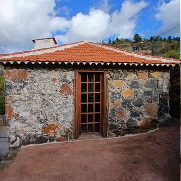 Dieses Bild zeigt ein kleines Steinhaus mit rotem Ziegeldach, einer Holztür mit Glasscheiben, vor einem hellen Himmel und einem hügeligen Hintergrund.