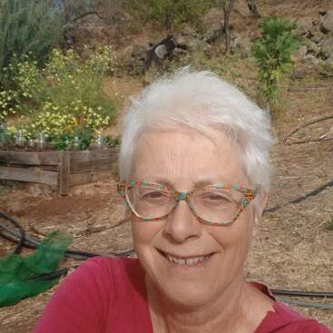 Eine Frau mit kurzen weißen Haaren und einer bunten Brille lächelt im Freien. Hinter ihr befinden sich ein Hochbeet, gelbe Blumen, Bäume, Felsen und ein strahlend blauer Himmel.