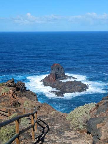 Un sentier côtier rocailleux descend vers un rocher volcanique spectaculaire entouré de vagues océaniques déferlantes, sous un ciel d'un bleu éclatant avec des nuages épars. Une scène magnifique et sereine.