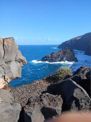 Des rochers massifs et sombres encadrent une vue côtière époustouflante où les vagues turquoise de l'océan se heurtent à des falaises escarpées sous un ciel bleu limpide, créant un paysage naturel spectaculaire.
